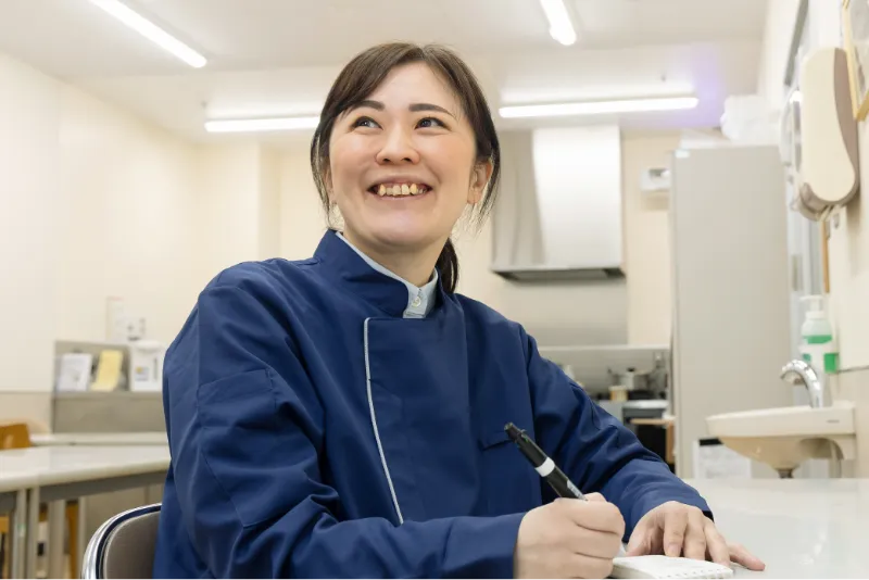 Y.Y.さんがバックヤードで椅子に座り、メモ帳に何かを記入している途中で顔をあげて、誰かと話しているような様子