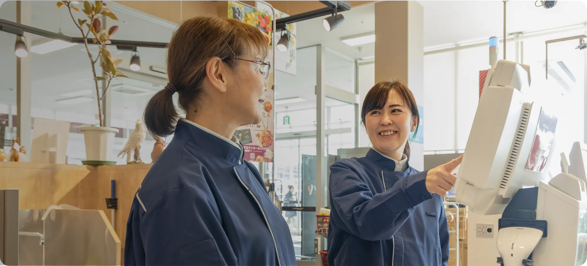 従業員二人がレジ前に立ち、一人がレジの画面を指さしながら、もう一人の従業員へ笑顔で話しかけている様子