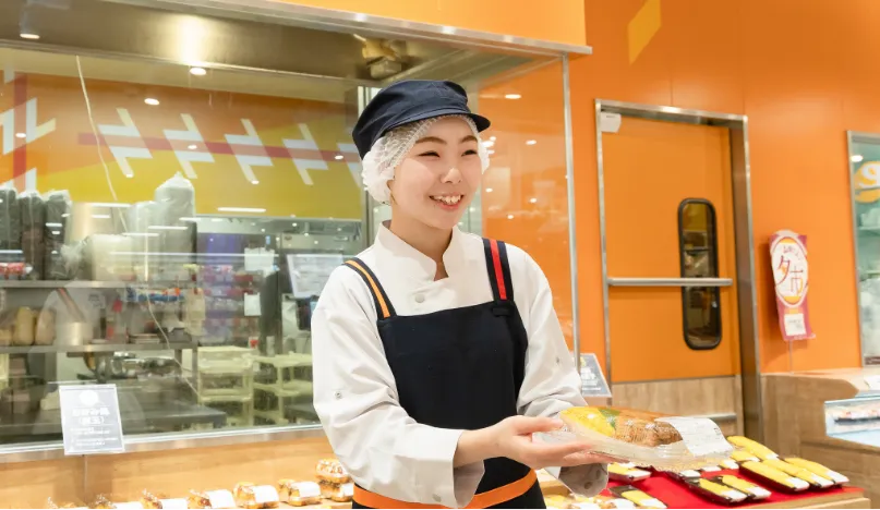 お惣菜売り場で、お惣菜のパックを持った従業員が、笑顔でお客様に商品をお渡ししようとしている様子