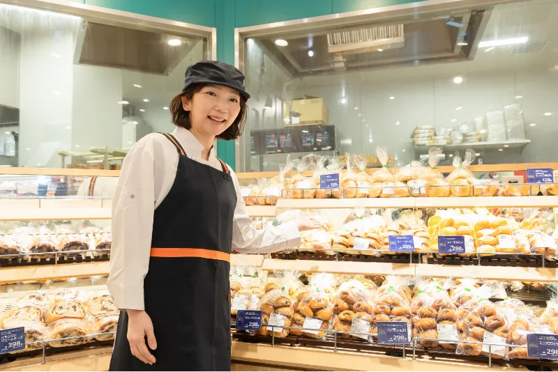 ベーカリーコーナーで、H.T.さんが手のひらを上に向け、パンをお客様に案内してお勧めする様子