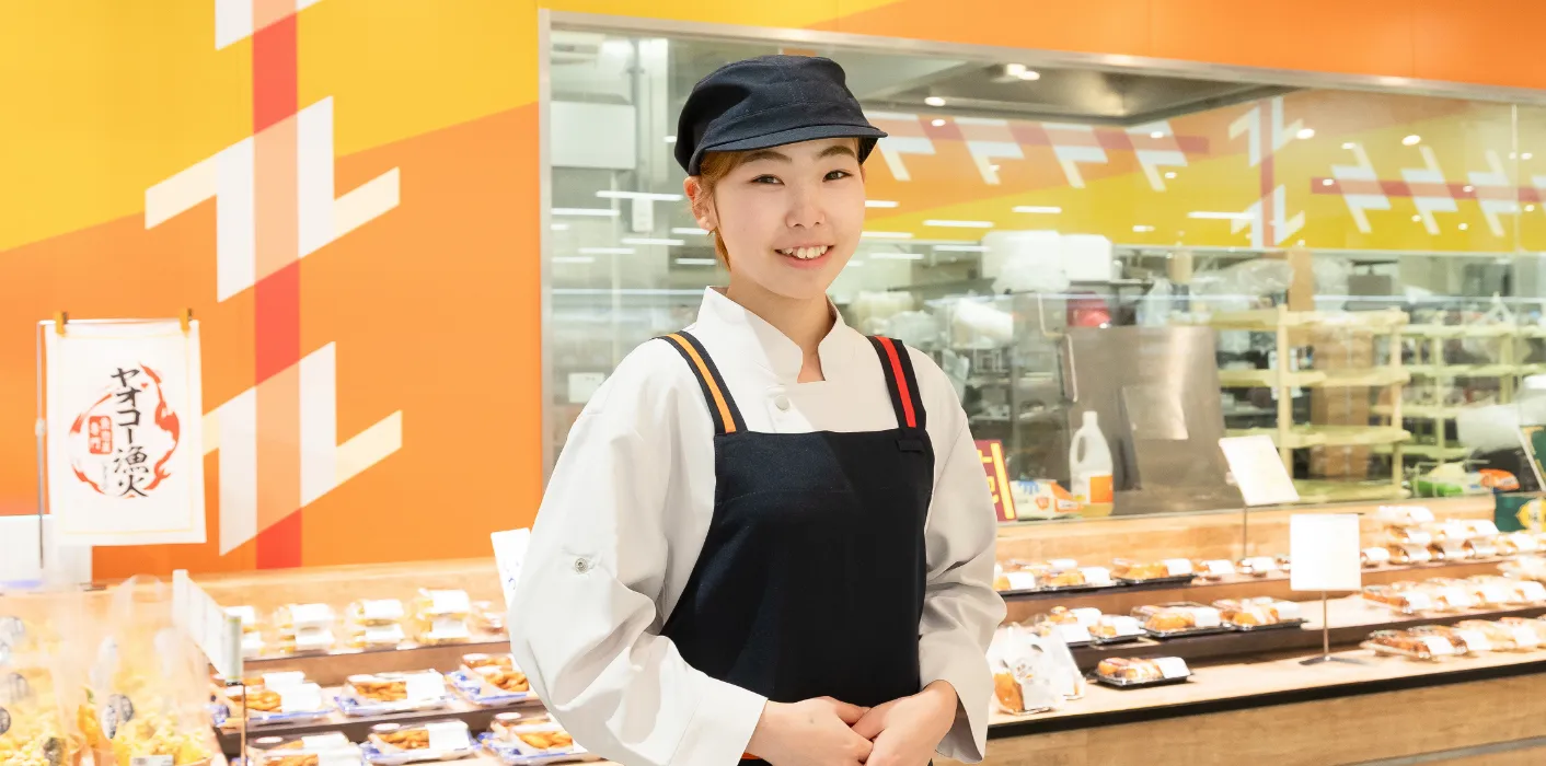 M.A.さんがお惣菜コーナーを背にして笑顔で立っている様子