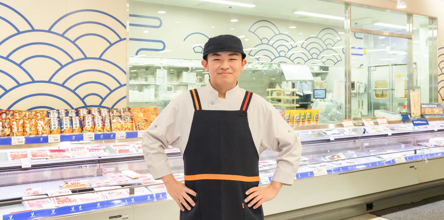 S.E.さんが鮮魚コーナーを背にして、腰に手を当て笑顔で立っている様子